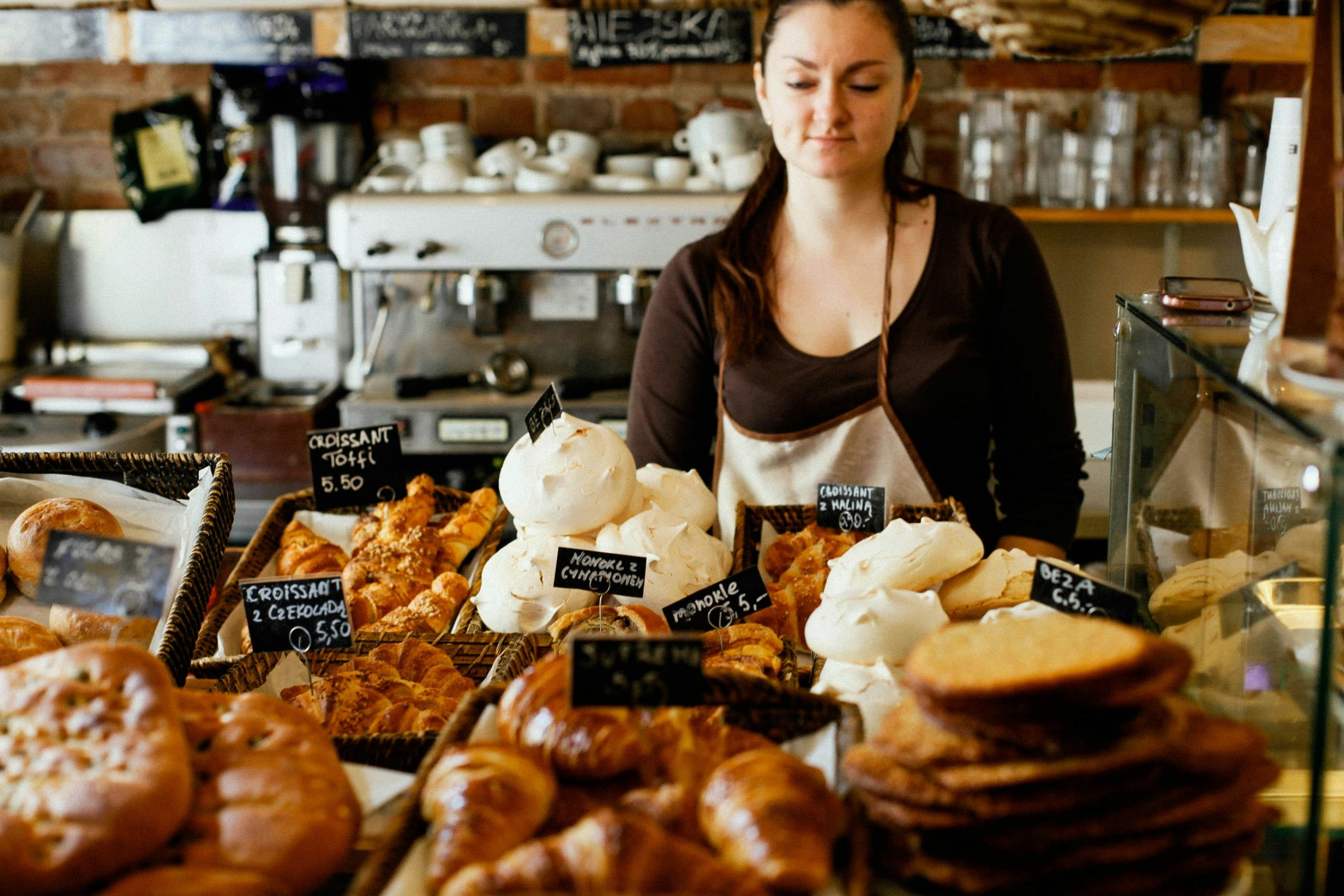 White woman in a brown shirt and cream apron standing behind a bakery counter with various baked goods on display