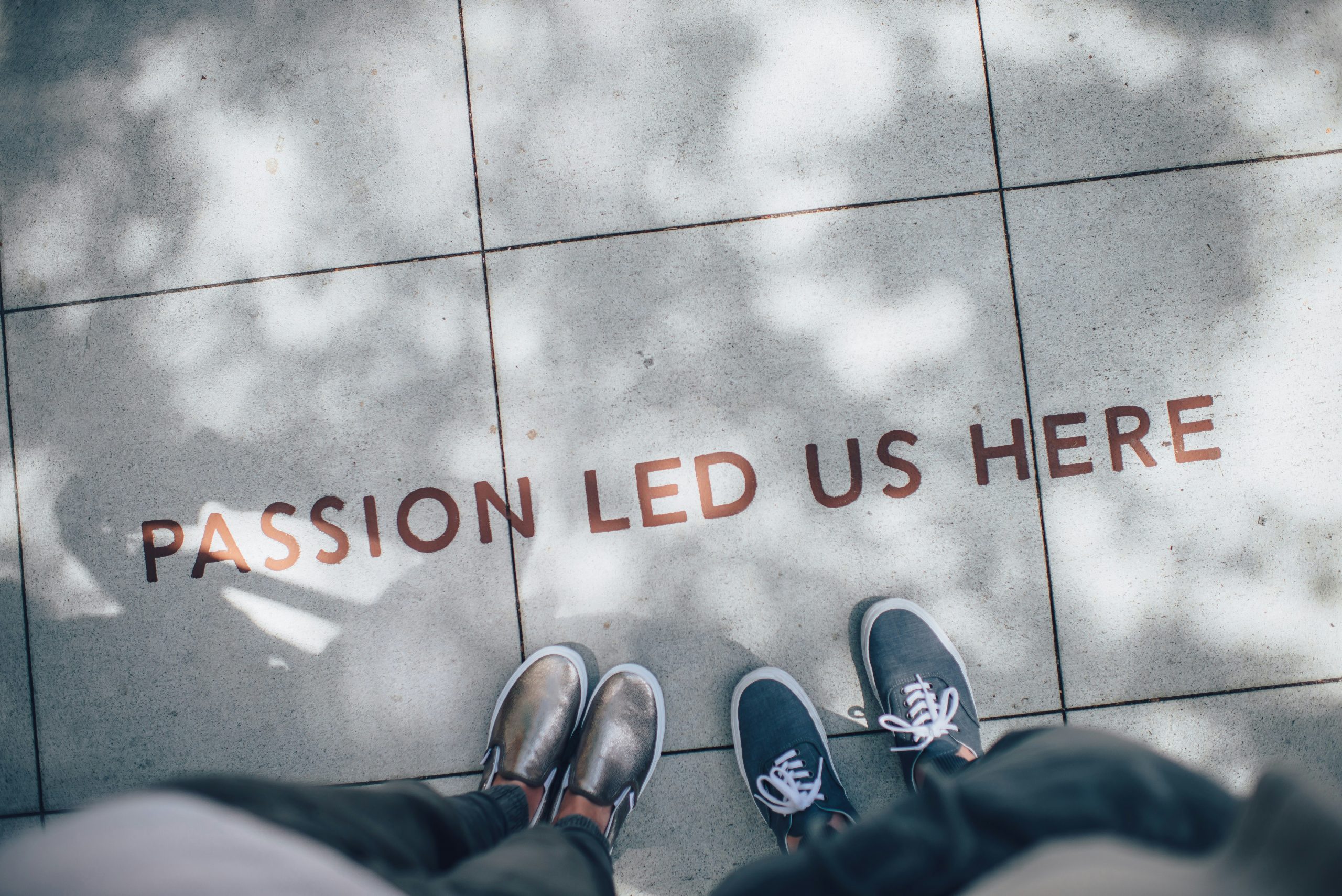 The phrase "Passion Led Us Here" on a sidewalk with two people standing by it (you see their shoes)