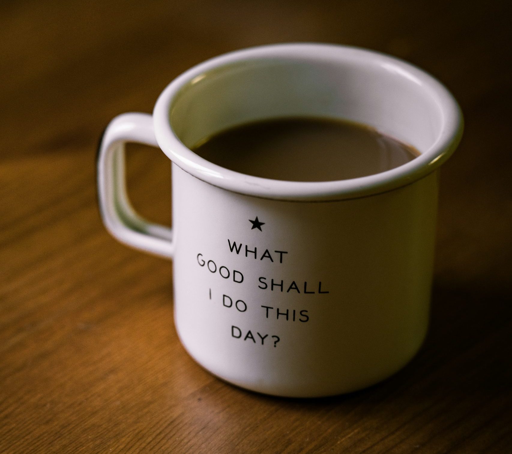 white ceramic mug full of coffee on a wood surface that reads "What Good Shall I Do This Day?"