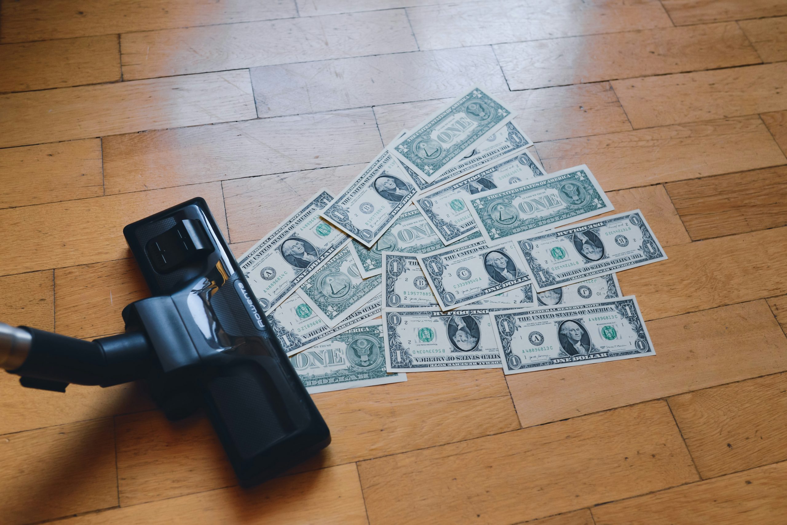 A black vacuum cleaner sucking up US dollar bills on a hardwood floor background